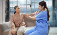 A care worker or nurse helping an older woman through an arm exercise, while both are sitting on a sofa in a living room