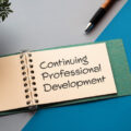 The words 'continuing professional development' written on the page of a pad which is sitting on a table alongside a pen and a plant