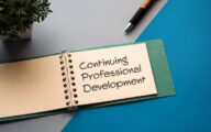 The words 'continuing professional development' written on the page of a pad which is sitting on a table alongside a pen and a plant