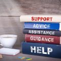 Pile of books on a wooden desk with the words 'advice', 'assistance', 'guidance' and 'help' on the spines