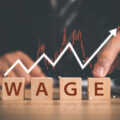 Blocks spelling out the word 'wage' with a line graph above it and a man in a suit pointing at it