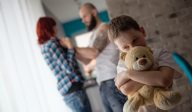 A child hugs a teddy bear in the foreground while in the background a person with a beard is holding someone with red hair threateningly