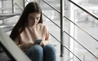 Upset teenage girl with smartphone sitting on stairs indoors.