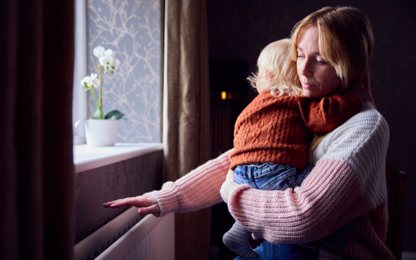 Mother With Son Trying To Keep Warm By Radiator At Home During Cost Of Living Energy Crisis