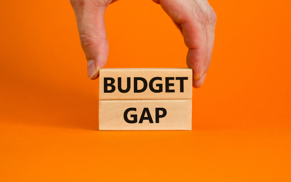 Person placing a wooden block with the word 'budget' written in black on it on top of a similar block with the word 'gap' written on it