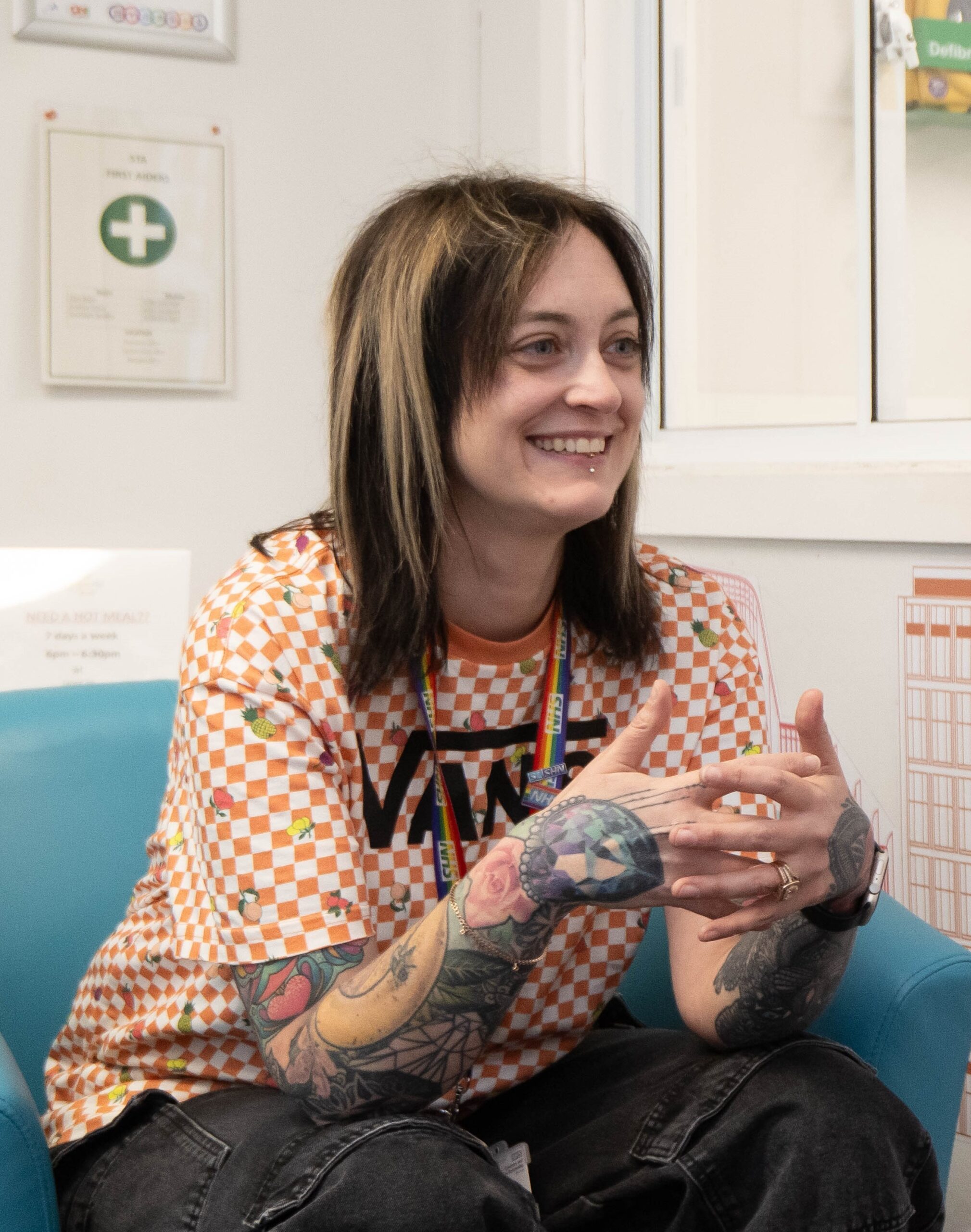 Sara Galvin sitting in a blue chair wearing a orange and white chequered Vans t-shirt and smiling.