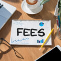 The word 'fees' written on a notepad in the middle of a desk, alongside a computer, phone and some pencils