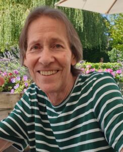Tony Inwood in 2023. He is wearing a stripy top and smiling at the camera. Behind him are flowers and green trees