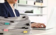 Stack of documents and woman working with laptop at table in office, closeup