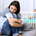 Young mother looking stressed in foreground with baby in cot in background
