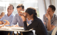 Indian woman laughing at funny joke eating pizza with diverse coworkers in office, friendly work team enjoying positive emotions and lunch together, happy colleagues staff group having fun at break