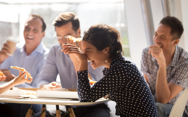 Indian woman laughing at funny joke eating pizza with diverse coworkers in office, friendly work team enjoying positive emotions and lunch together, happy colleagues staff group having fun at break